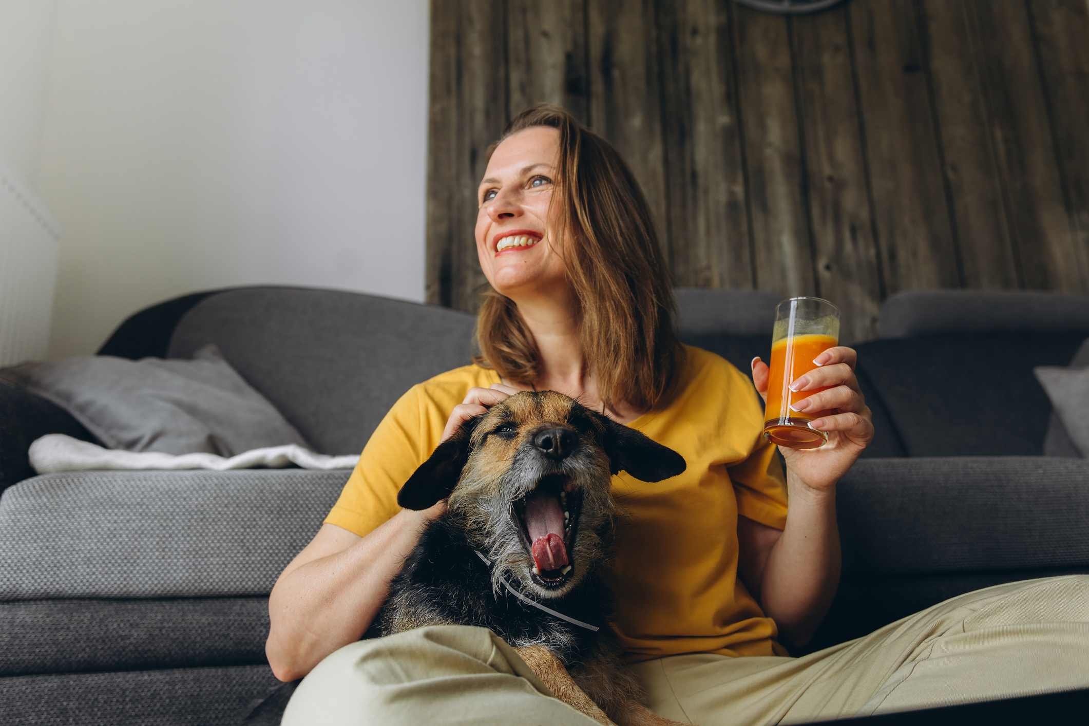 Smiling woman in a yellow shirt relaxing at home with her dog while holding a glass of fresh orange juice