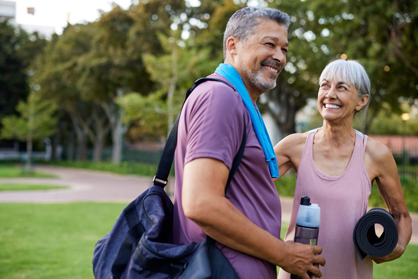 Smiling, active middle-aged couple with gym bags, a water bottle, and a yoga mat enjoying time outdoors in a park